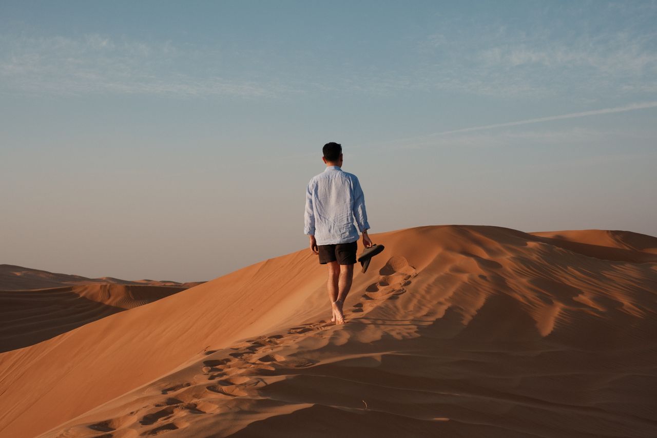 A man walks barefoot on vast desert sand dunes, creating footprints under a clear sky.